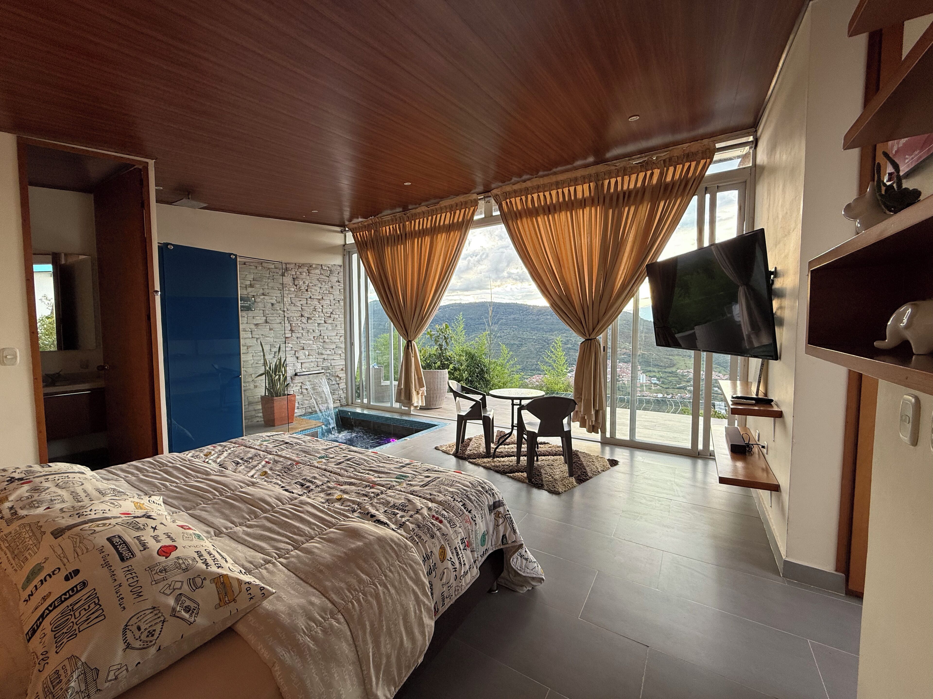A bedroom with a large bed, a TV, a small table with chairs, a potted plant, and a view of the outdoors.