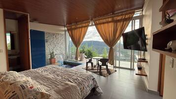 A bedroom with a large bed, a TV, a small table with chairs, a potted plant, and a view of the outdoors.