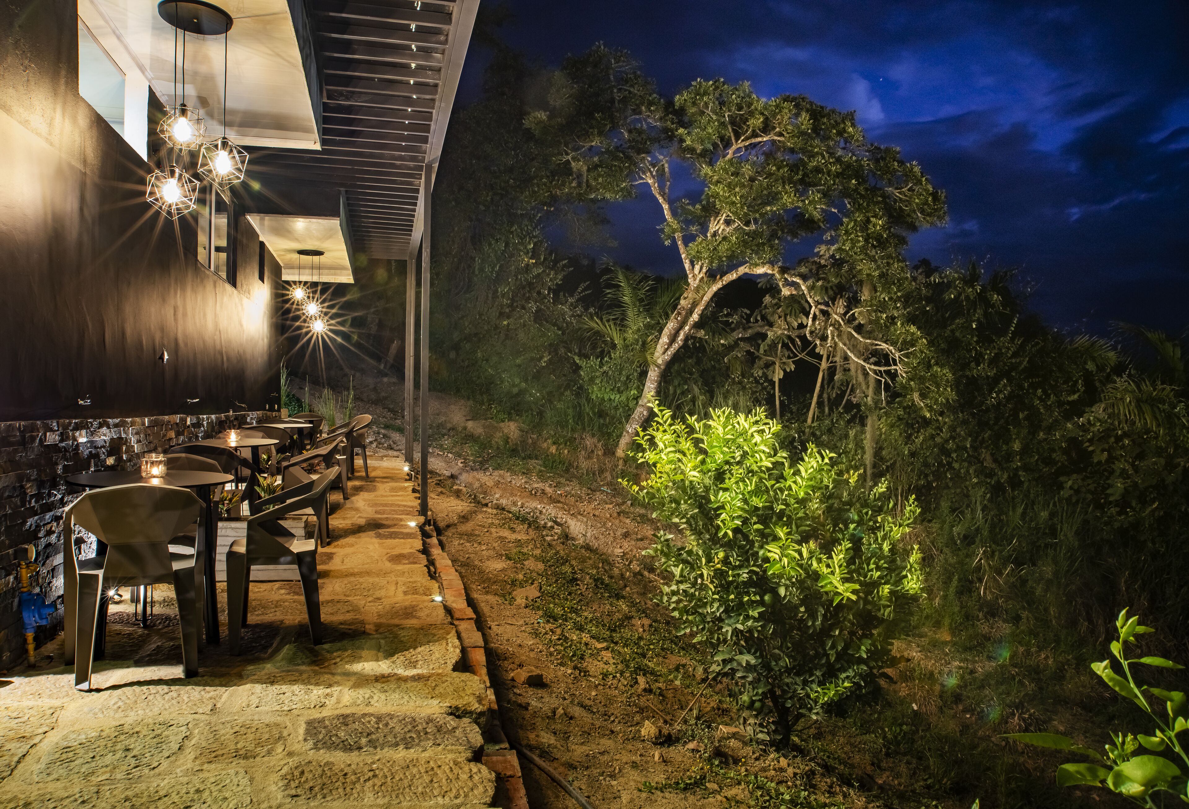 An outdoor dining area with stone flooring, tables, and chairs, surrounded by greenery and a tree.
