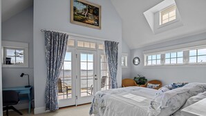 Une chambre avec un grand lit, un bureau et une chaise. Elle comporte deux fenêtres et une porte qui donne sur un balcon.