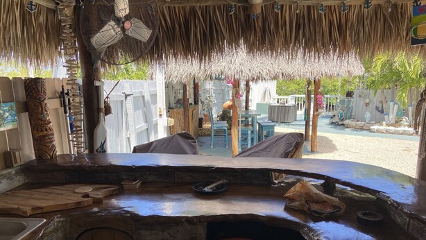 A rustic outdoor kitchen area with a thatched roof, wooden furniture, and a large fan.
