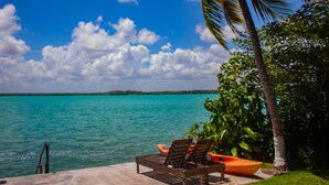 A beachside view with a wooden lounge chair and an orange kayak, palm trees, and turquoise water. - Propiedad a Orilla de Laguna (Huay-Pix)