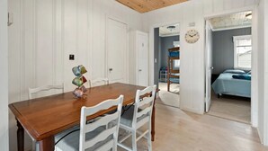 Un coin repas avec une table en bois et des chaises blanches, une horloge au mur et une vue sur une chambre à coucher avec un lit et une fenêtre.