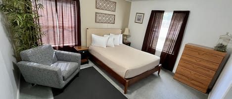 Une chambre à coucher comprenant un lit, une chaise, une commode, une table de chevet, une lampe, une plante et une fenêtre avec des rideaux.