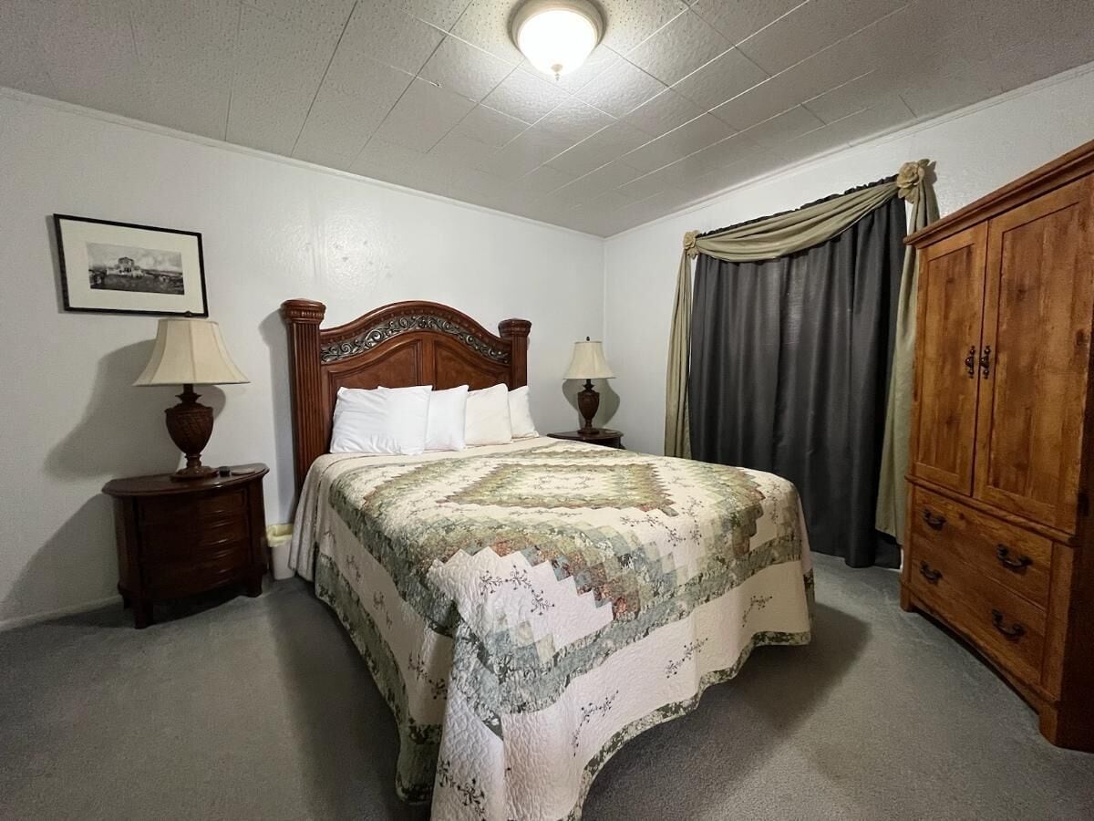A bedroom with a bed, two nightstands, a wooden wardrobe, a window with curtains, and a framed picture on the wall.