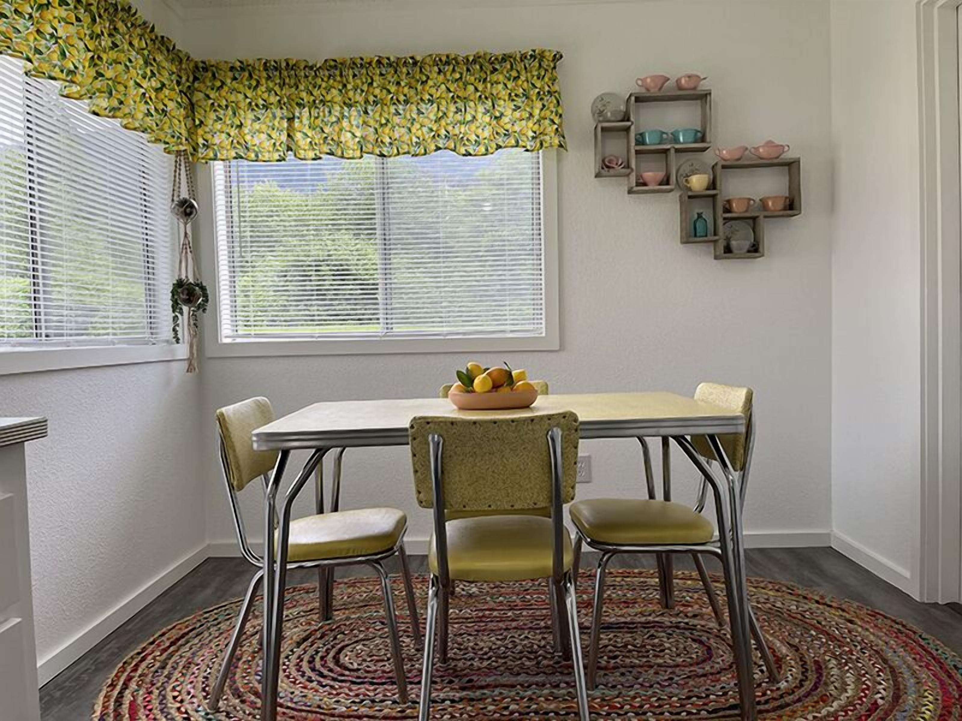 A dining area with a table, chairs, a patterned rug, a window with blinds, and a wall with shelves.