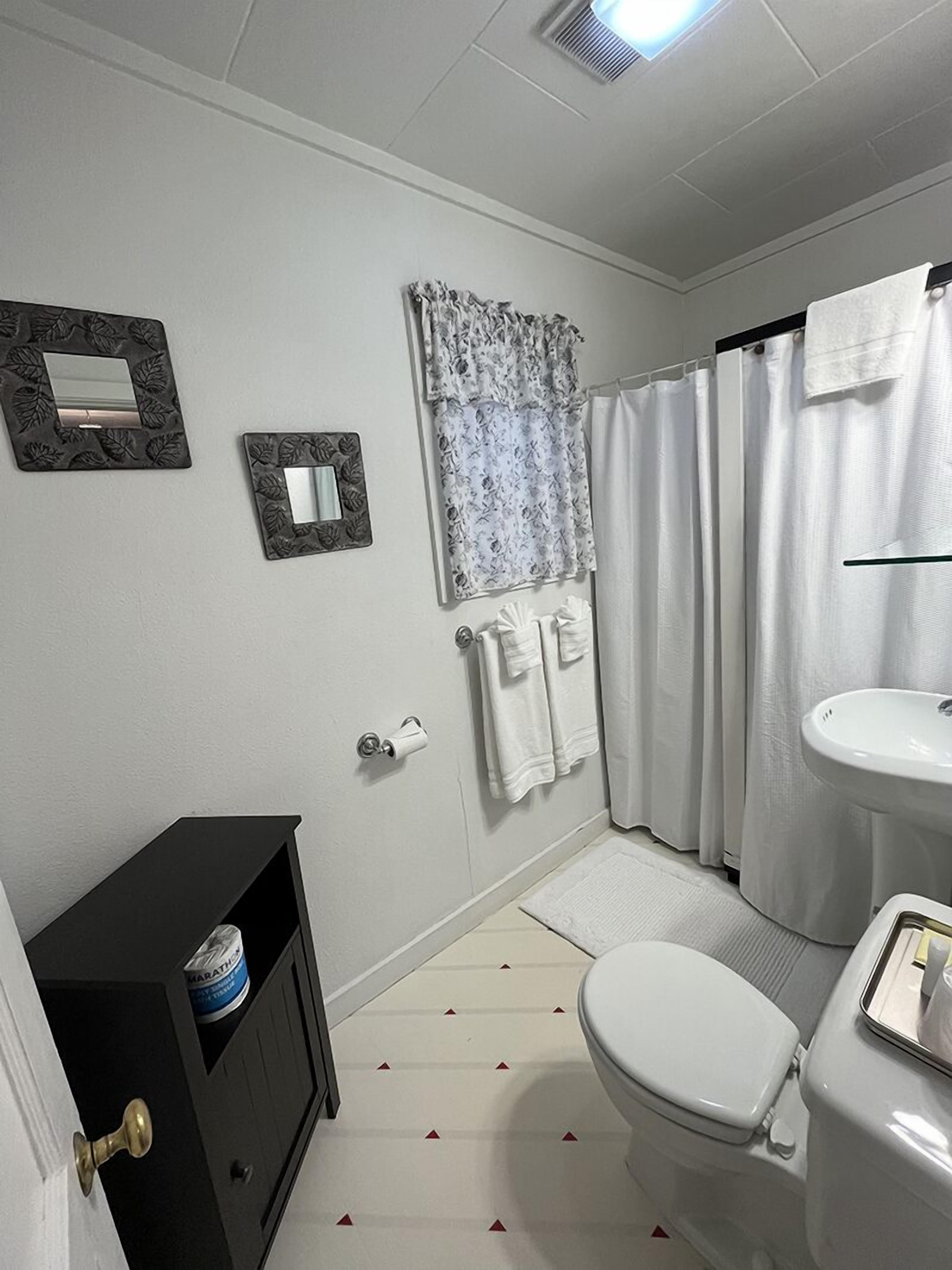 Et badeværelse med toilet, håndvask, bruseforhæng, håndklæder og et lille skab.