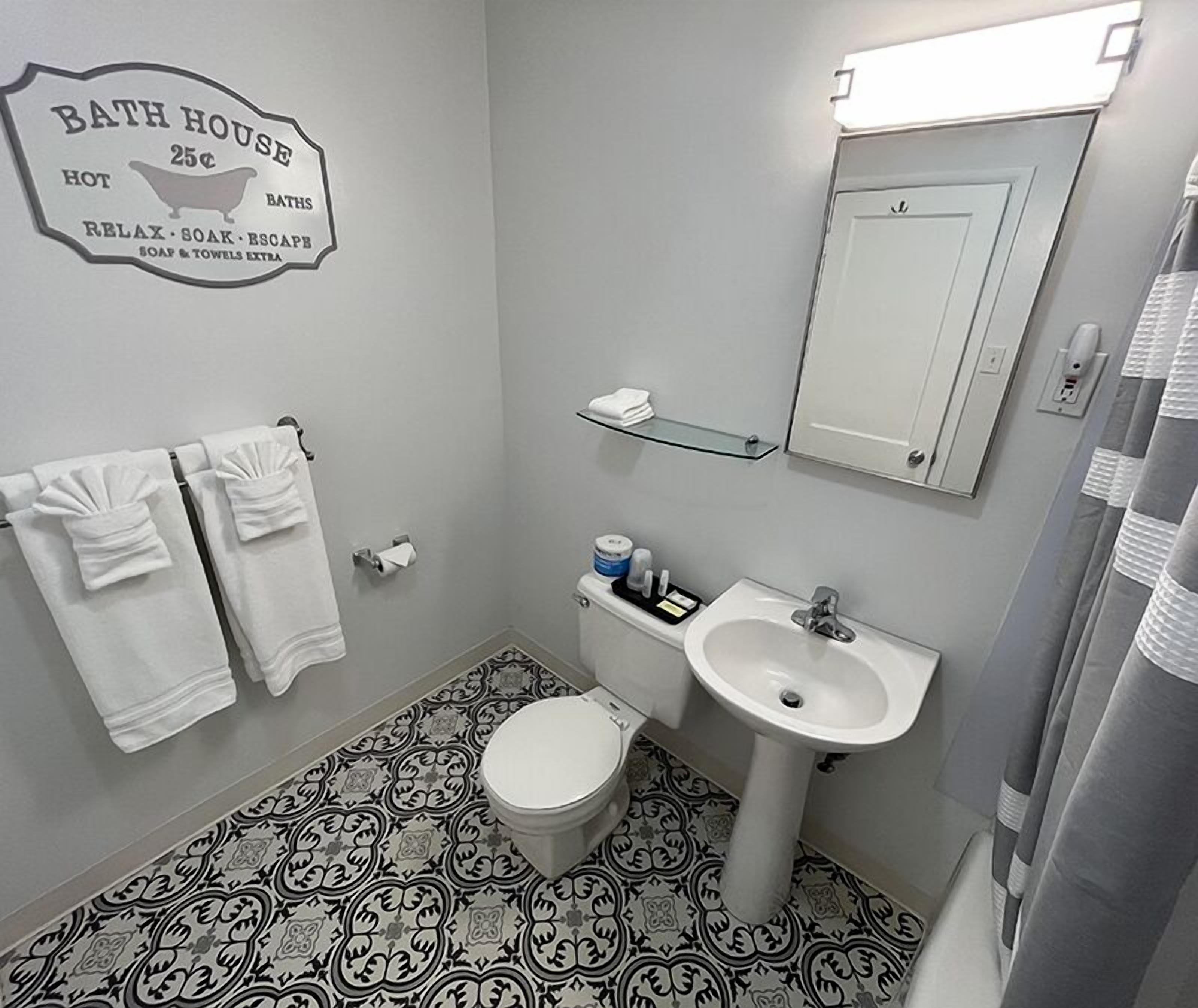 A bathroom with a toilet, sink, and towel rack. There's a sign on the wall advertising 'Bath House 25c' with services like hot baths, relax, soak, escape, and extra soap and towels.