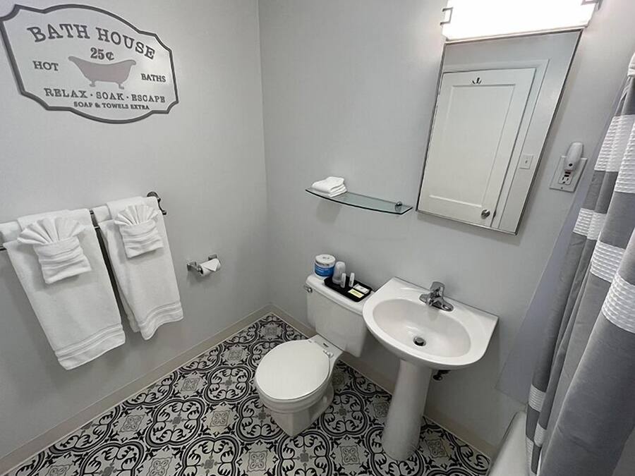 A bathroom with a toilet, sink, and towel rack. There's a sign on the wall advertising 'Bath House 25c' with services like hot baths, relax, soak, escape, and extra soap and towels.