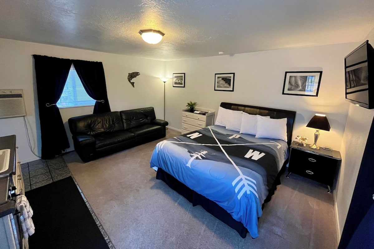 Un dormitorio con cama, sofá de cuero negro, mesita de noche con lámpara, calentador de pared y ventana con cortinas.