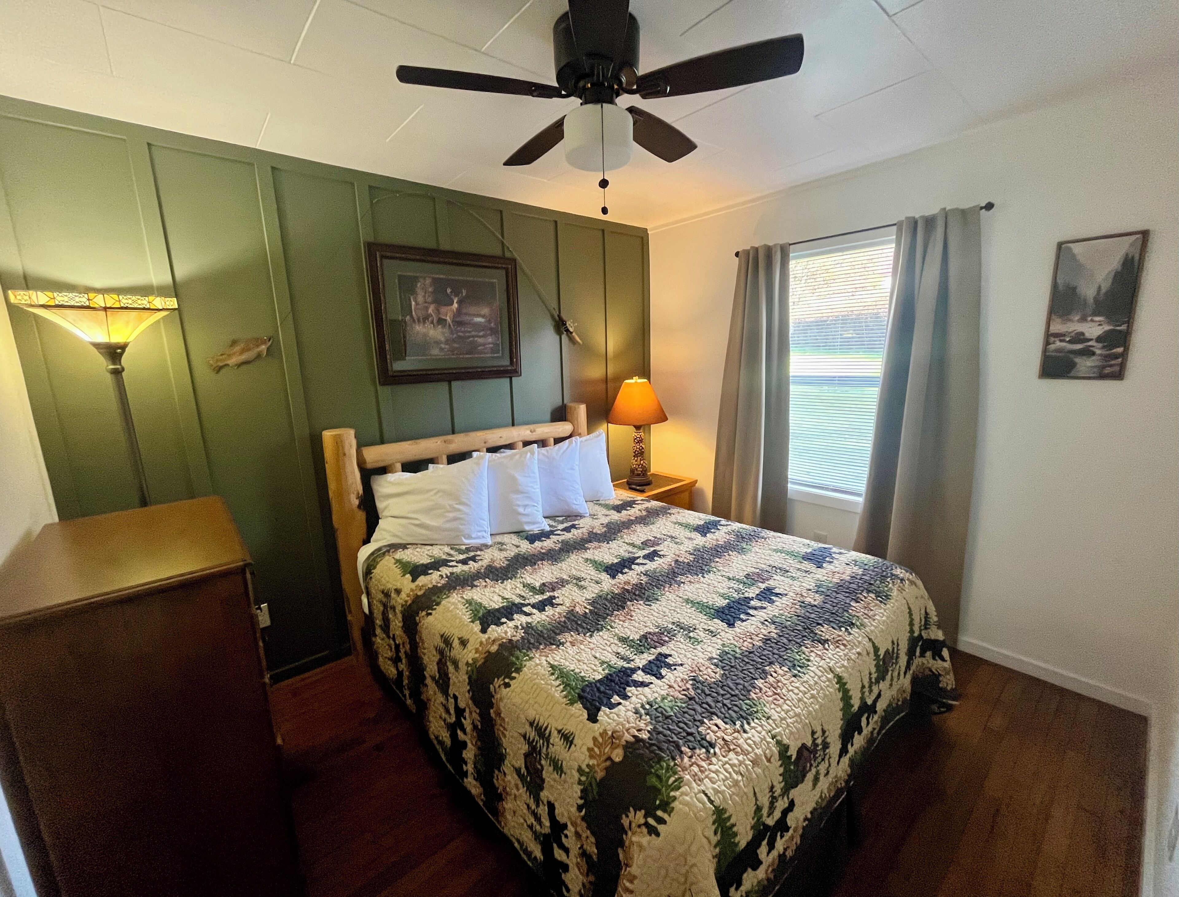 A bedroom with a bed, a ceiling fan, a nightstand, a lamp, a framed picture, and a window with curtains.