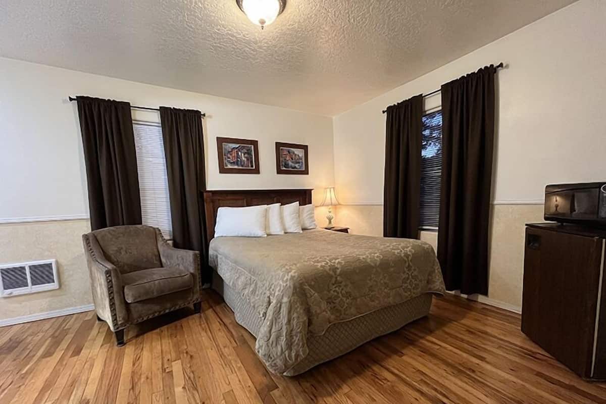 Habitación de hotel con piso de madera, cama con cabecera, una silla, mesita de noche con lámpara, televisor y dos cuadros en la pared.