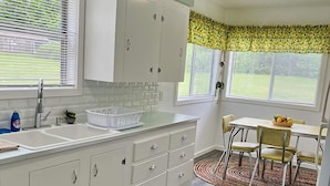 A kitchen with white cabinets, a white countertop, a backsplash, and a window with a yellow leaf-patterned valance. - Cascade Motel (Cascade Locks)