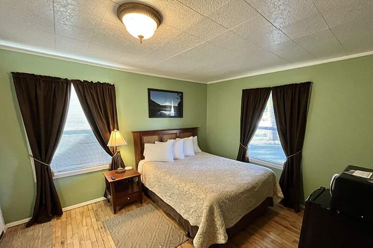Habitación de hotel con una cama, una mesita de noche, una lámpara, un cuadro en la pared y una ventana con cortinas.