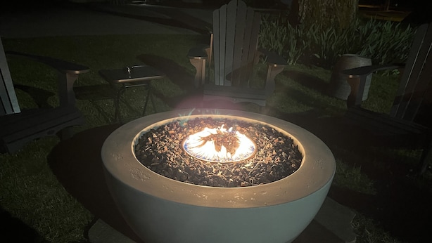 A circular outdoor fire pit with a visible flame, surrounded by a paved area with chairs and a wooden fence in the background. - Cascade Motel (Cascade Locks)