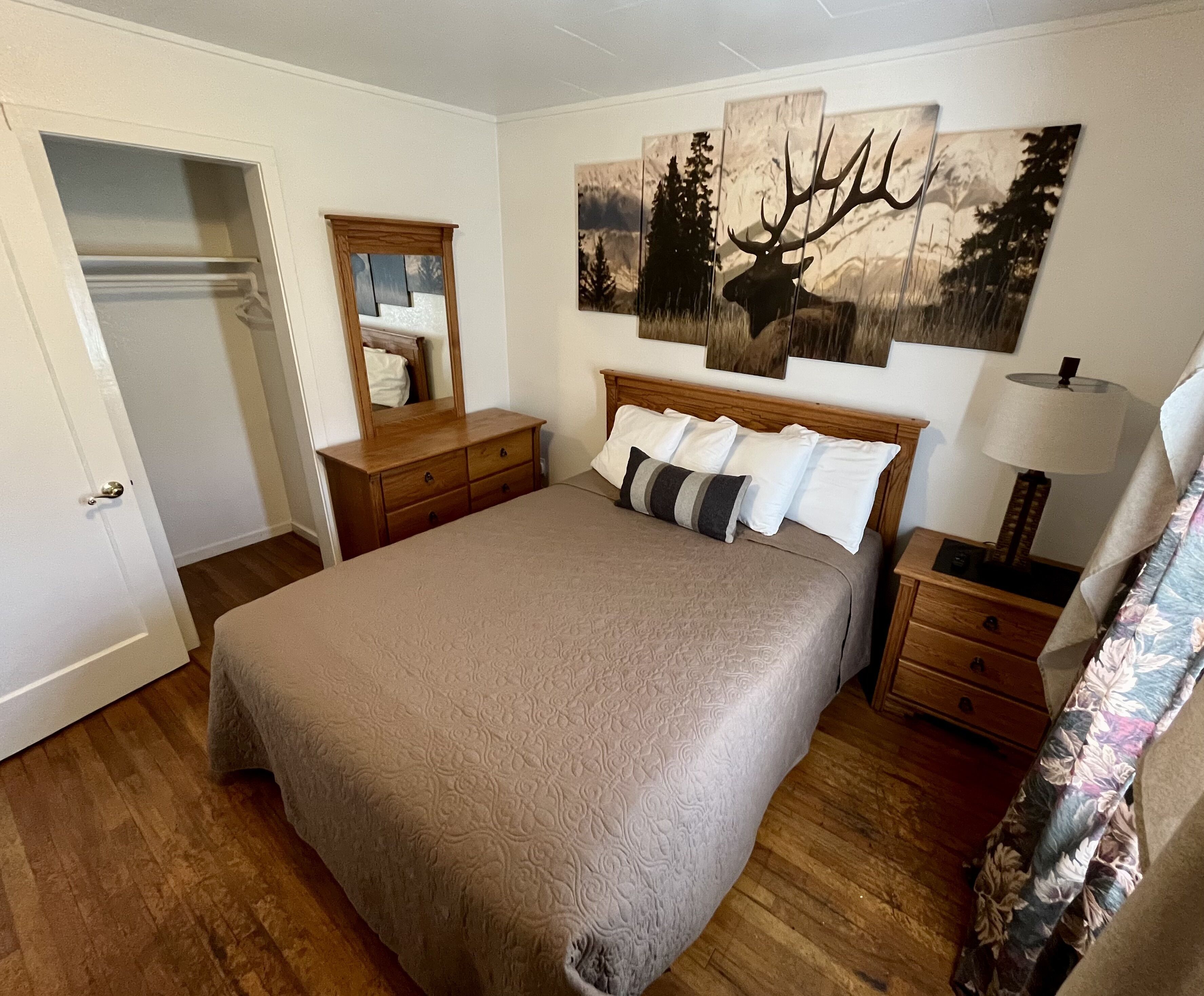 A bedroom with a large bed, wooden headboard, a nightstand with a lamp, a dresser, a mirror, and a painting of a deer on the wall.
