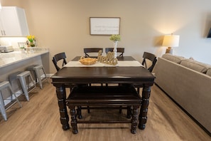 A dining area with a dark wooden table, two chairs, a bench, and a 'hello' sign. There is a framed quote on the wall, a vase with flowers, and a lamp on the right.