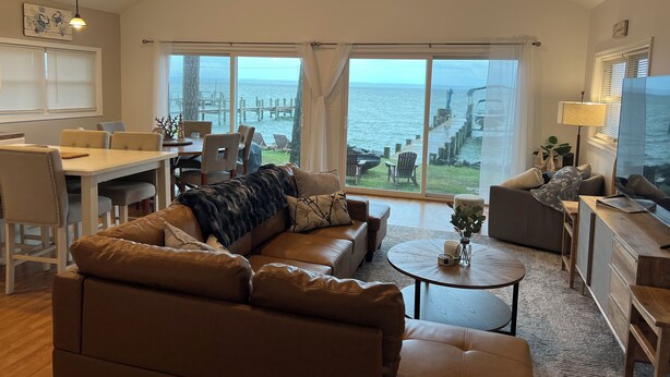 A spacious living room with a large window overlooking a dock and water, a ceiling fan, and a dining area with a table and chairs.