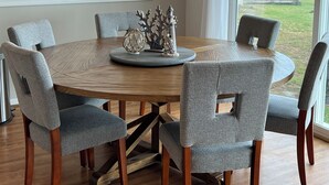 A dining area with a round wooden table, grey upholstered chairs, and a view of the outdoors through large windows.