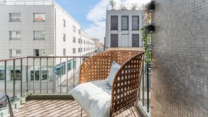 Un balcon doté d'un lit en osier, d'une rambarde en verre et offrant une vue sur les bâtiments voisins.