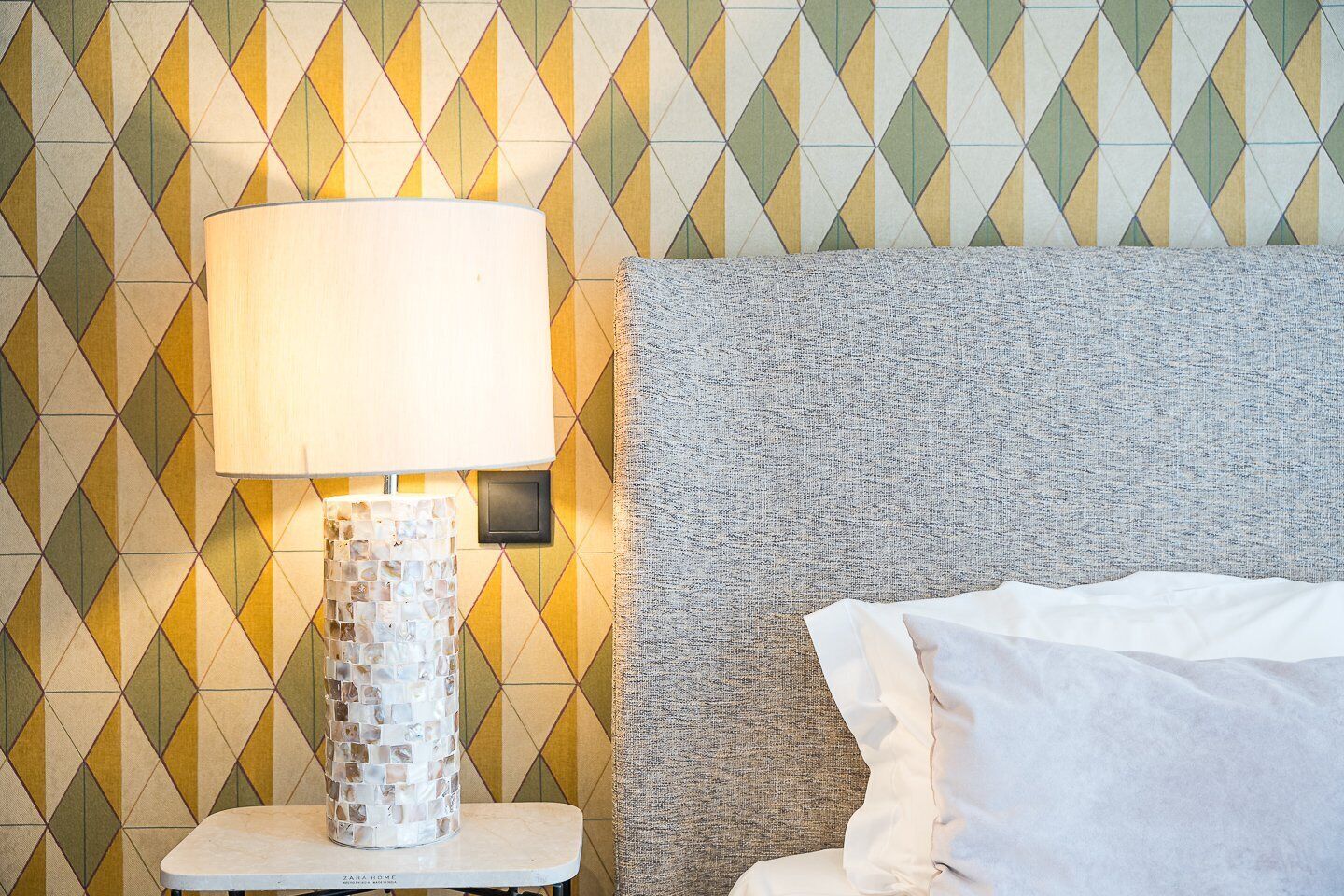 Uma cama com roupa de cama branca, cabeceira cinza texturizada e uma luminária com base em mosaico sobre um criado-mudo branco.
