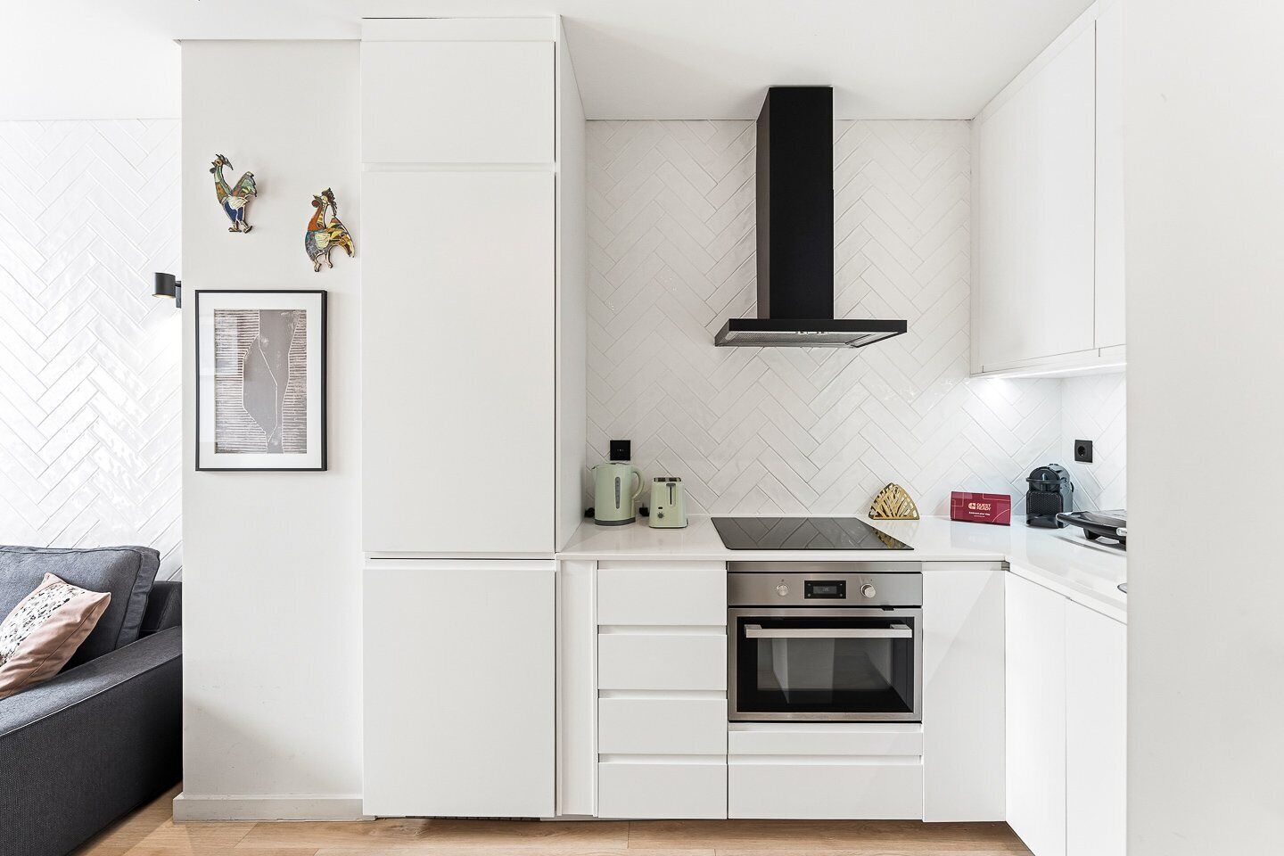 A modern kitchen with white cabinetry, a black range hood, a built-in oven, and a herringbone-patterned backsplash.