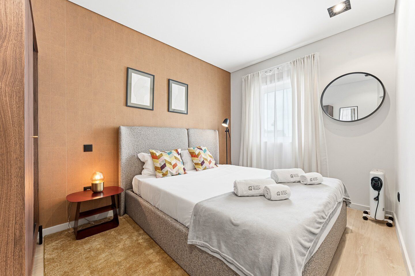 A modern hotel room with a large bed, a bedside table with a lamp, a round mirror, and a wall-mounted TV.