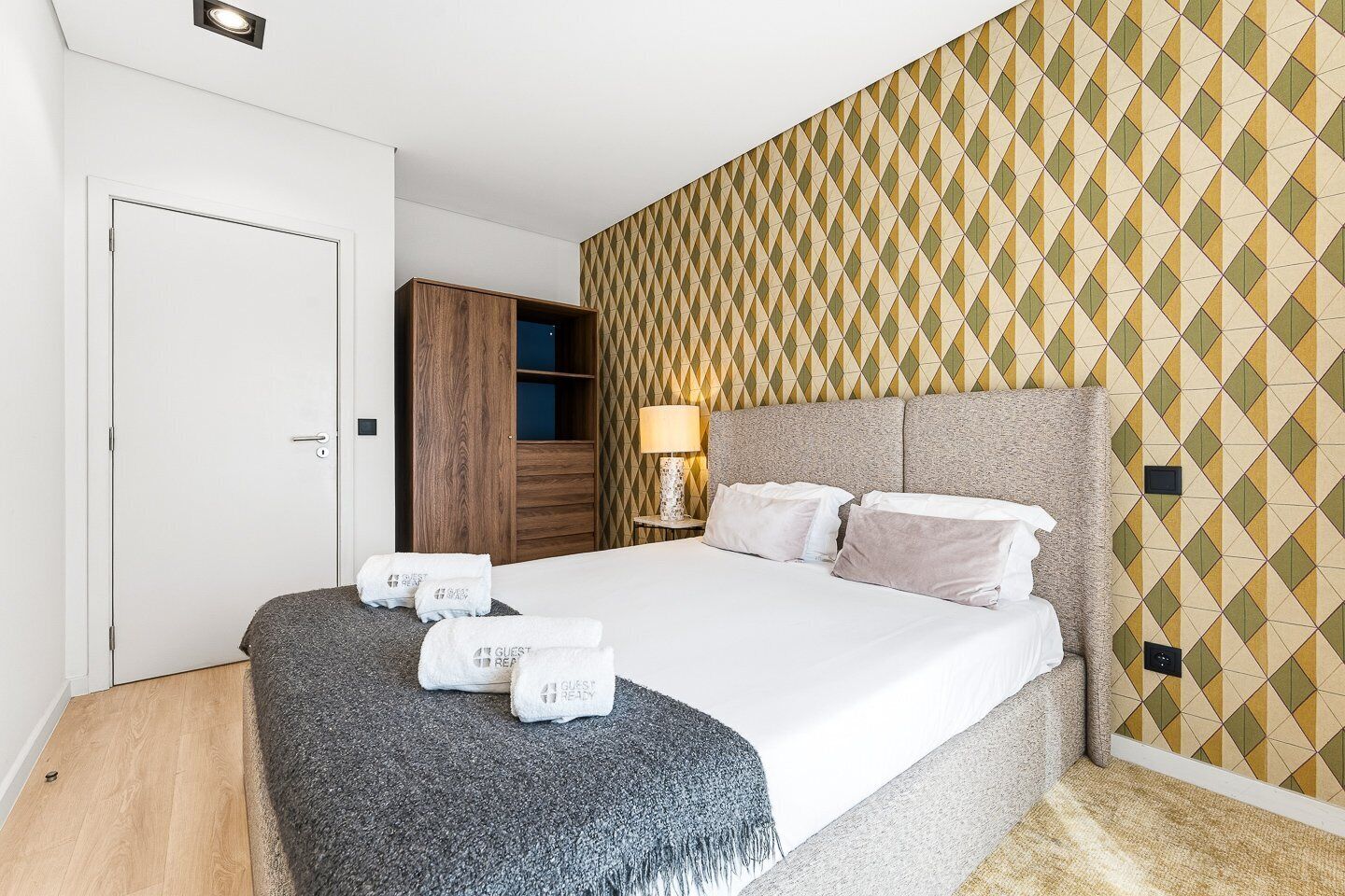 Quarto de hotel com cama, guarda-roupa, mesa de cabeceira com abajur e parede estampada.
