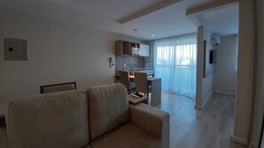 Een moderne woonkamer met een bank, een eethoek met stoelen, een keuken met een magnetron en een groot raam met gordijnen.
