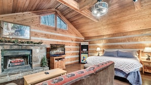 A log cabin interior with a stone fireplace, a bed with a blue blanket, and a television mounted on the wall. - Secluded Mountain Getaway - Spring Sale (Sevierville)
