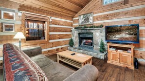 A cozy log cabin interior with a stone fireplace, a television, and a large window overlooking the forest. - Secluded Mountain Getaway - Spring Sale (Sevierville)