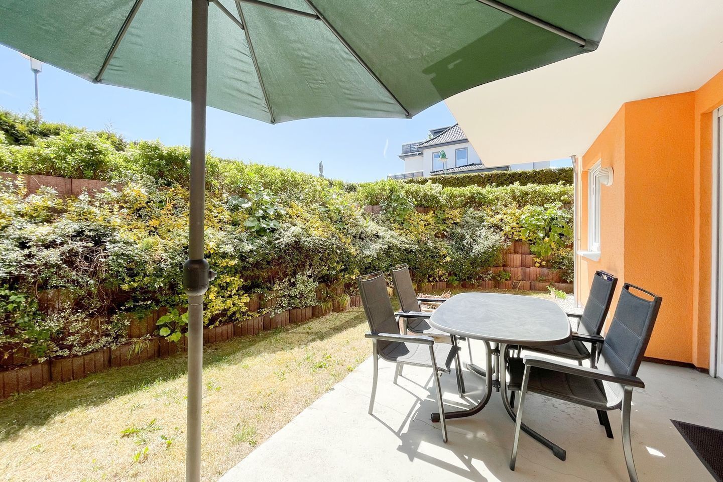 Une terrasse avec une table et des chaises, entourée d’une haie et d’une clôture en bois.