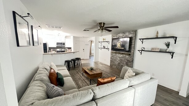 A modern living room with a sectional sofa, a coffee table, and a TV mounted on a stone wall. There are two black bar stools and a ceiling fan. - Home Away from Home (Haines City)