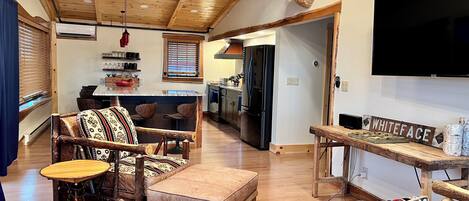 A rustic interior with a wooden ceiling, a mounted deer head, a bar area with stools, and a television on the wall.