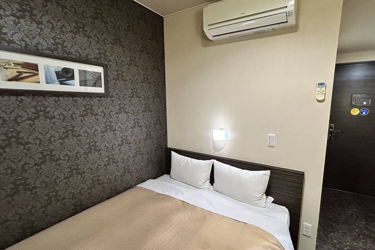 A hotel room with a bed, a wall-mounted air conditioner, a framed picture, and a door with a lock.