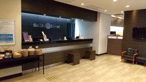 A reception area with a dark wood reception desk, a sign reading "Smile Hotel", and a seating area with two chairs. - Smile Hotel Kokura (Kitakyushu)