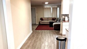 Un couloir au plancher de bois mène à un salon où se trouvent un canapé beige et un tapis rouge. Une poubelle est visible sur la droite.