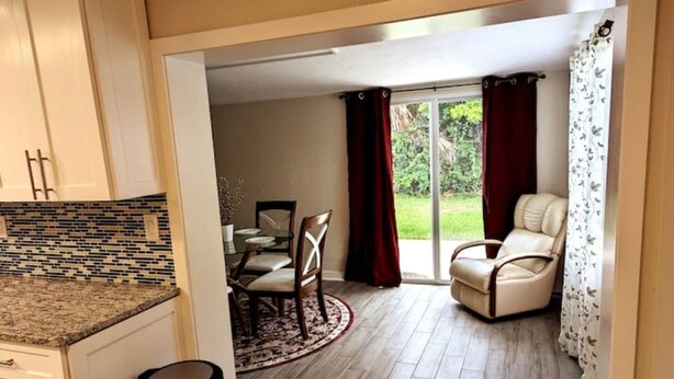 Une cuisine avec un mur recouvert de carreaux, un coin repas avec une table et des chaises, et une terrasse avec un fauteuil et des rideaux.