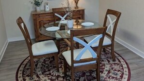 Un coin repas comprenant une table ronde en verre, six chaises en bois avec des coussins blancs, un tapis à motifs, une lampe fixée au mur et un tableau au-dessus de la table.