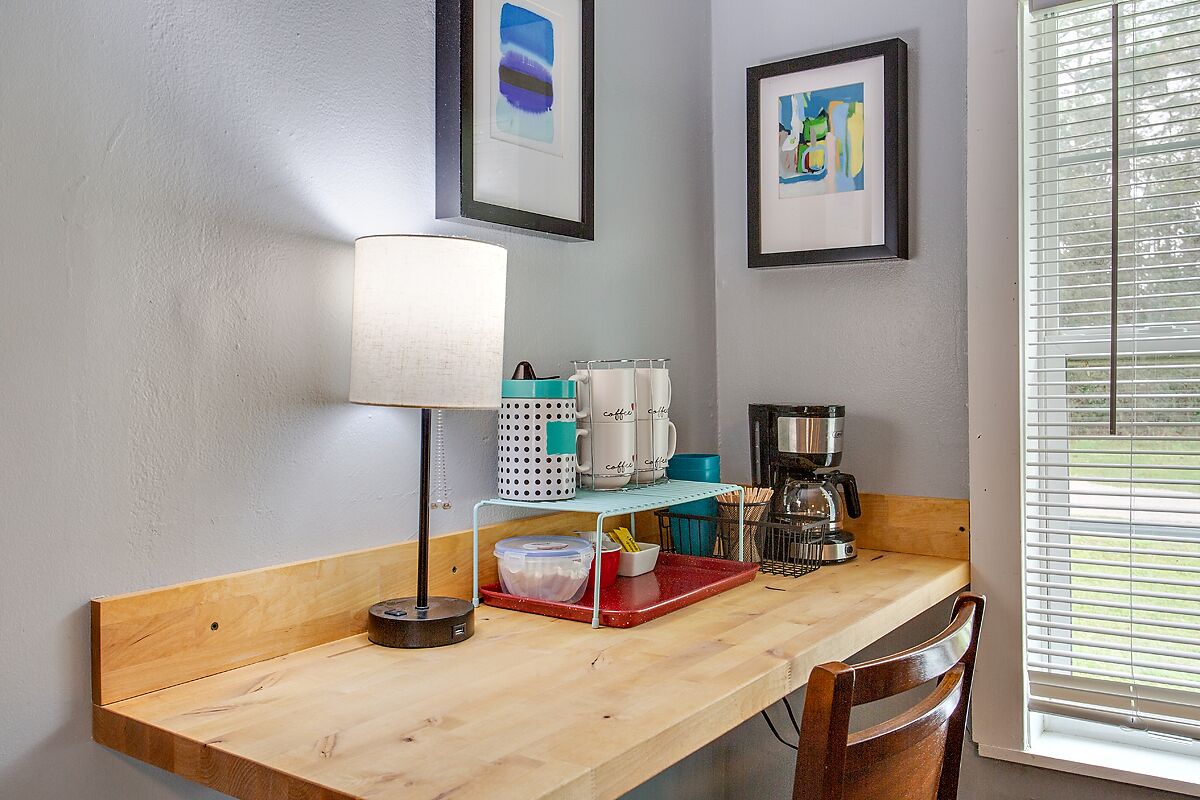 A small kitchenette with a wooden countertop, a coffee maker, a kettle, and a coffee cup. There is a framed picture on the wall and a window with blinds.