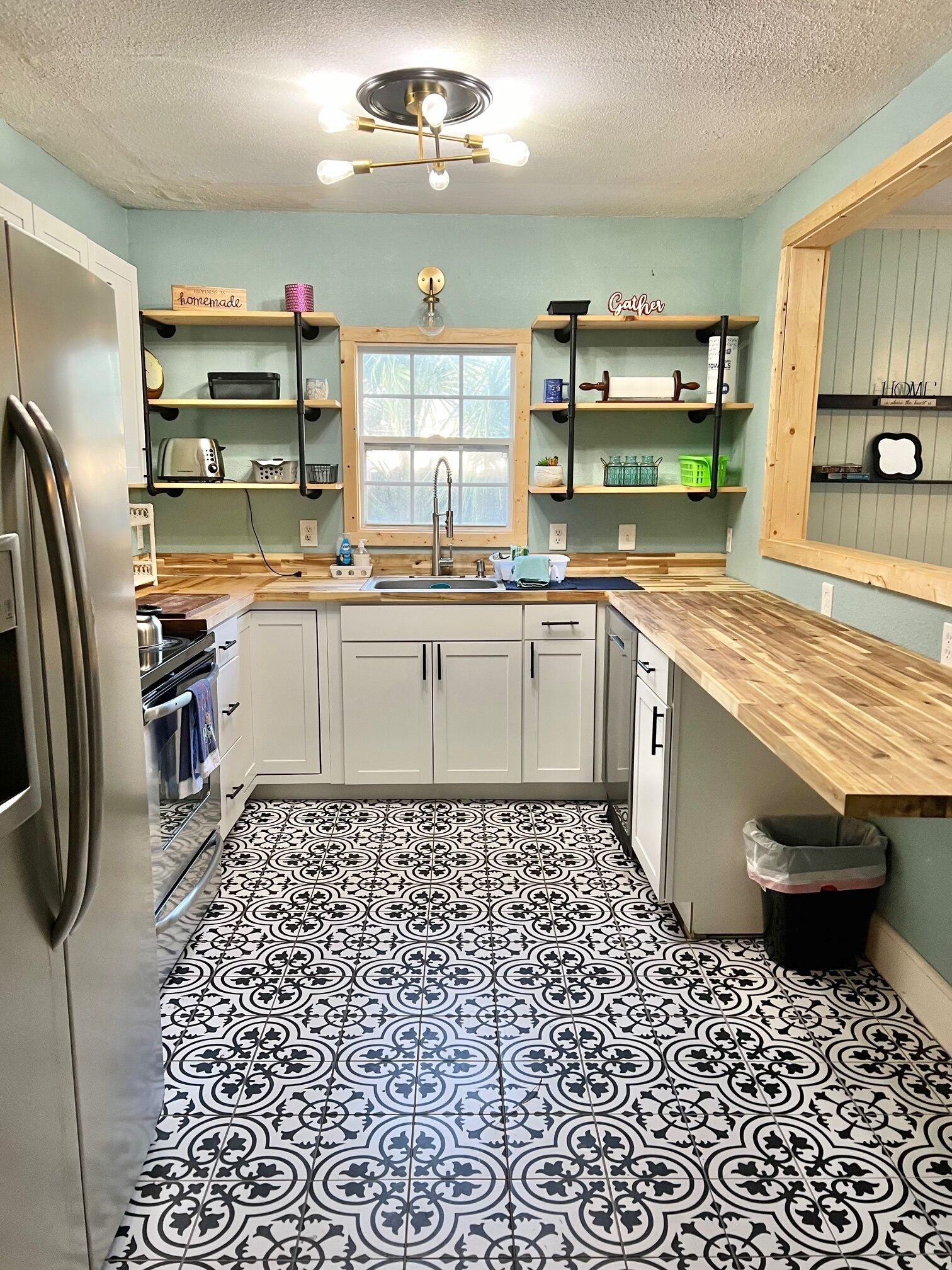 A kitchen with a black and white patterned floor, a stainless steel refrigerator, a wooden countertop, and a window above the sink.