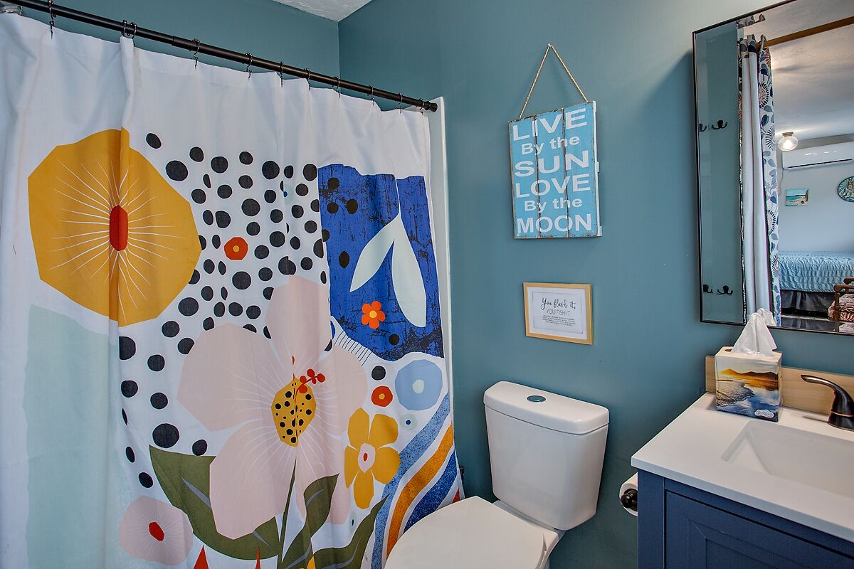 A bathroom with a shower curtain featuring floral and abstract designs, a toilet, a sink with a mirror, and a wall with a motivational quote.