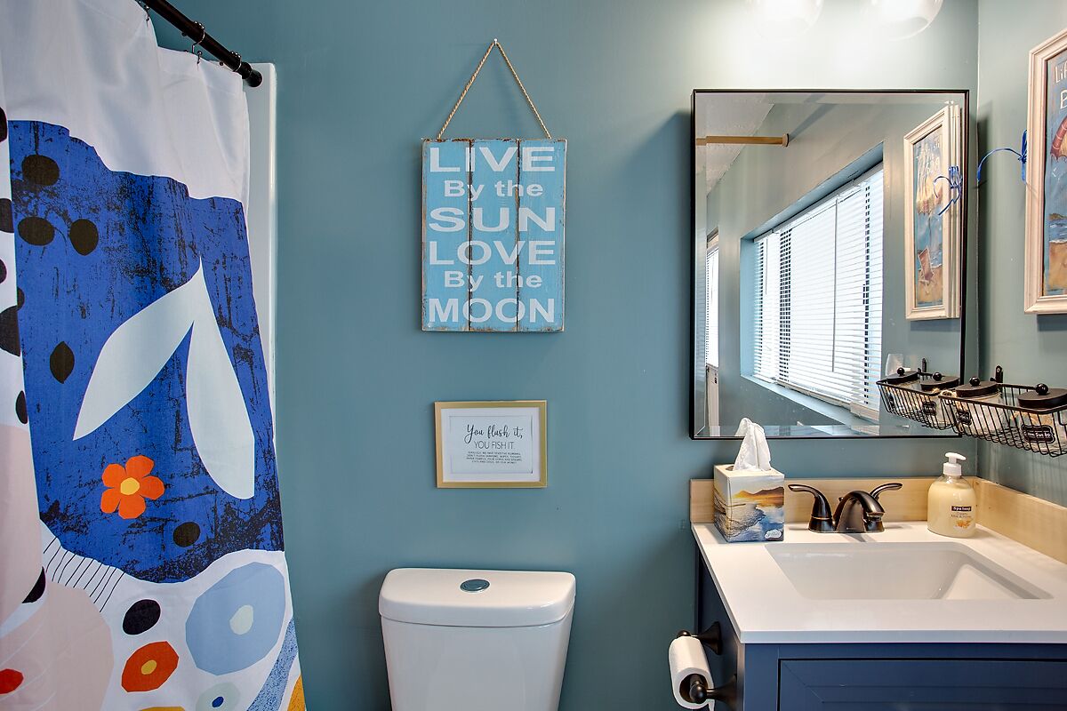 A bathroom with a blue shower curtain, a toilet, a sink with a mirror, and a framed sign saying "LIVE By the SUN Love By the MOON".