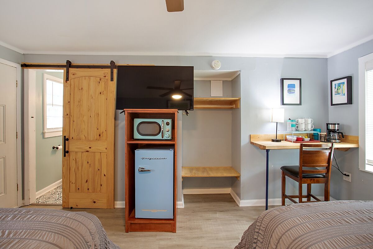 A room with two beds, a wooden door, a small kitchenette with a microwave, a ceiling fan, a desk with a chair, and a window.