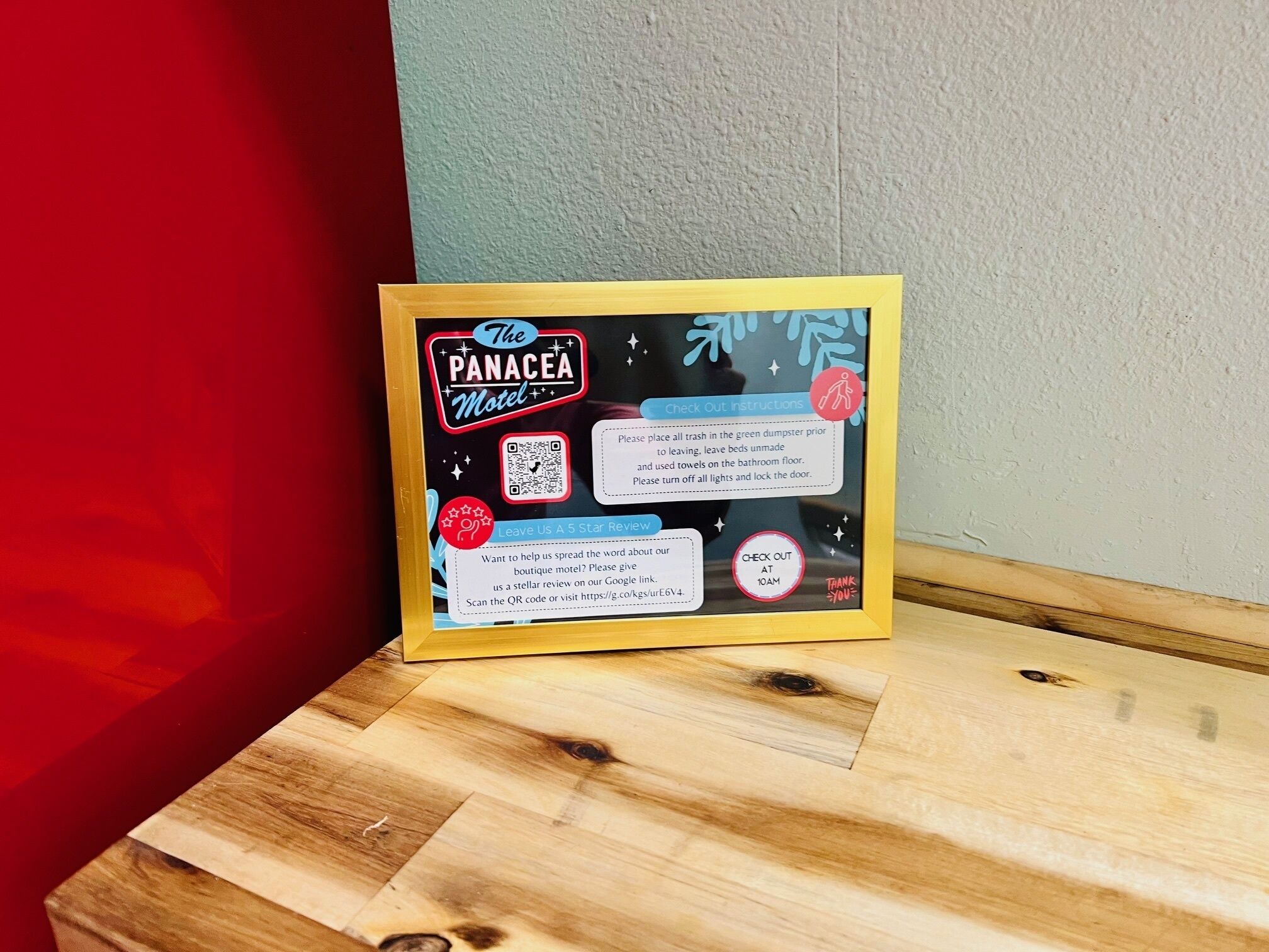 A framed sign for The Panacea Motel with check-out instructions and a QR code for reviews.
