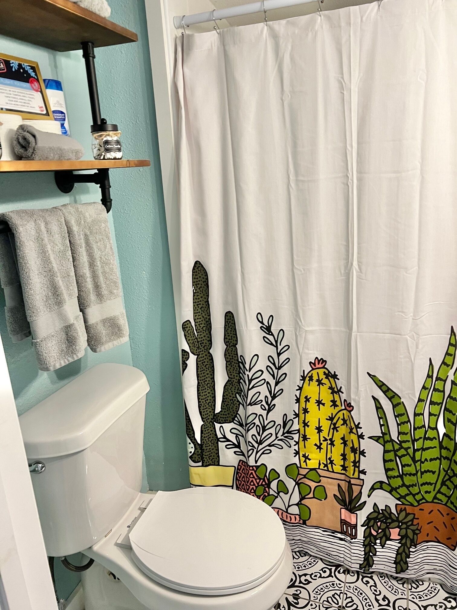 A bathroom with a toilet, a shelf with towels, and a shower curtain with a cactus design.