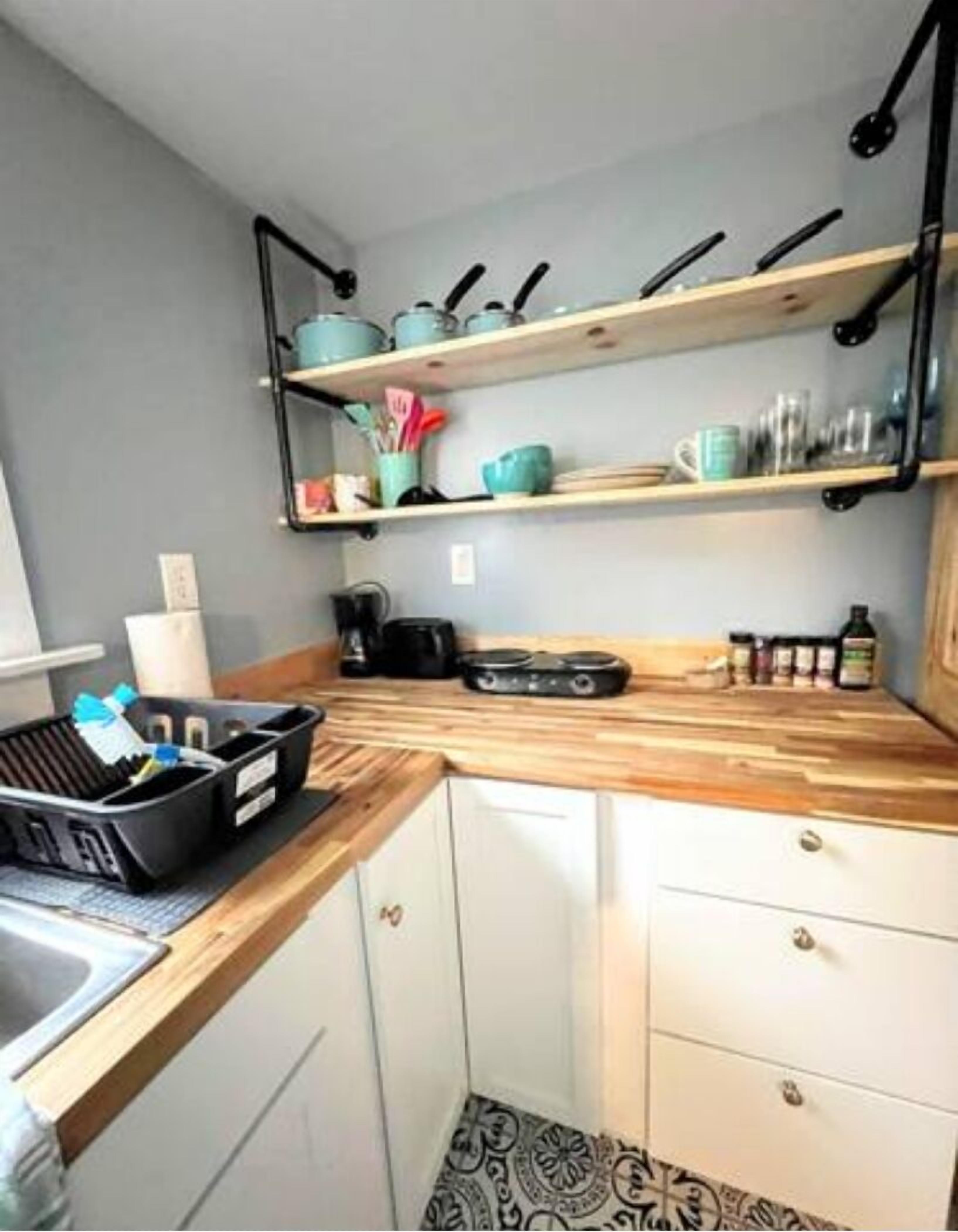 A compact kitchen with white cabinets, a wooden countertop, and two shelves holding kitchenware.
