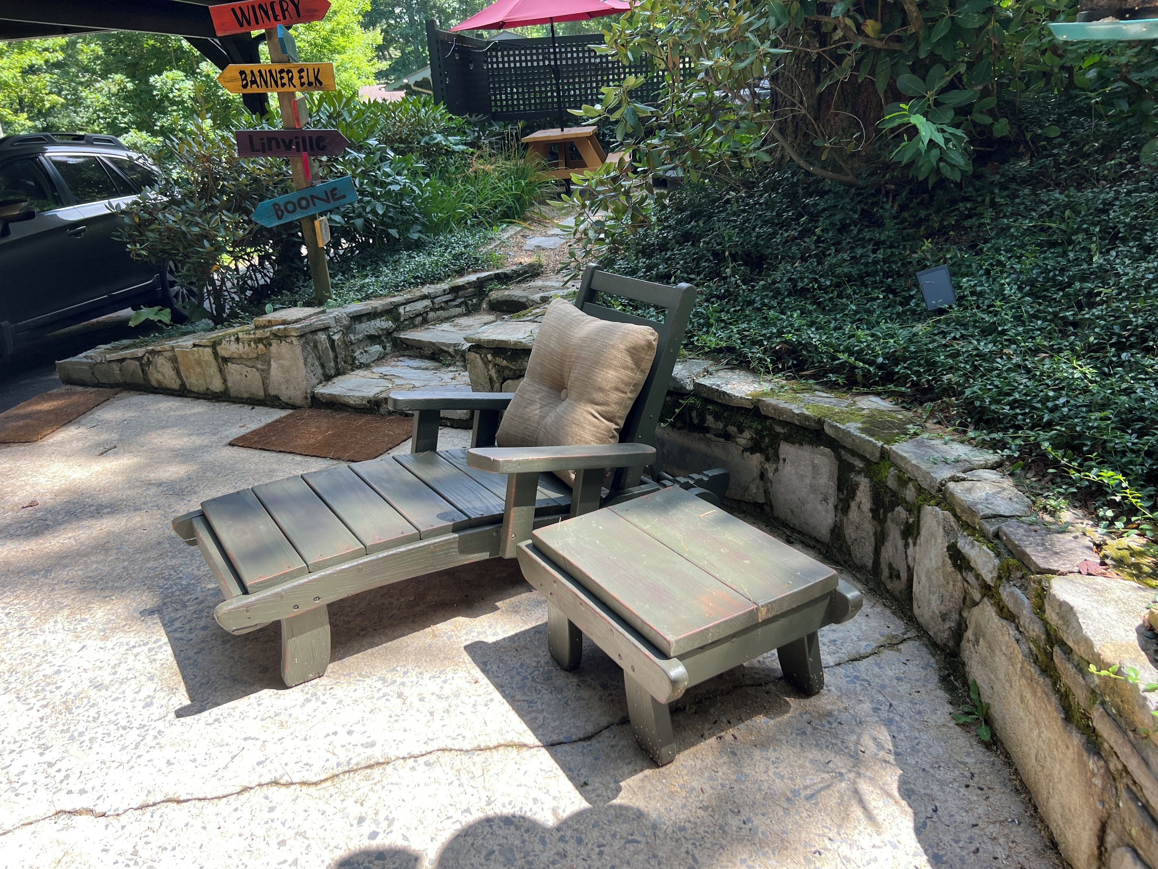 Una zona patio con una poltrona di legno e un tavolino, circondata da un muro in pietra e vegetazione.