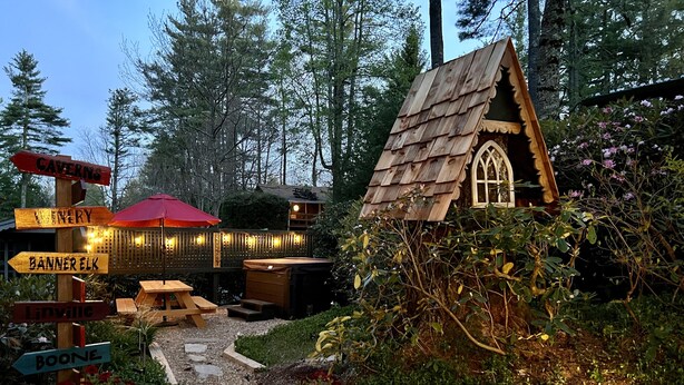 Een rustieke houten hut met schuin dak en raam, omgeven door weelderige begroeiing en een houten terras.