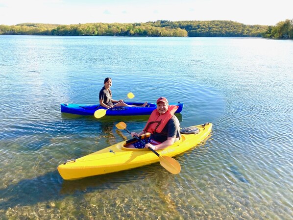 Two people kayaking on a calm lake with clear water, surrounded by trees and hills in the background.